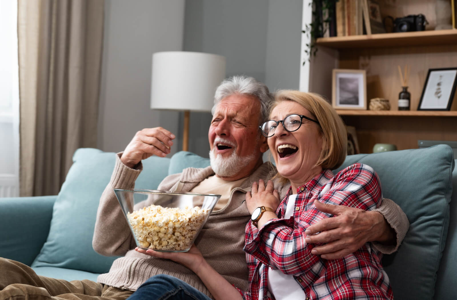 An older retired couple laughing on the couch with popcorn while watching a movie together.