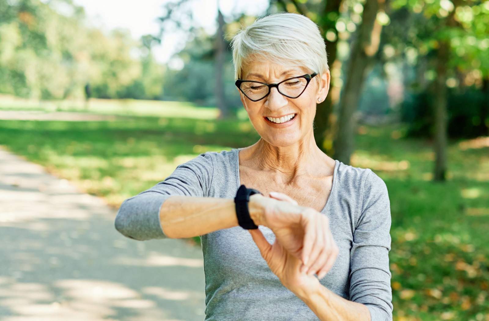 Smiling senior wearing glasses checking smartwatch on walking path surrounded by trees and sunlight