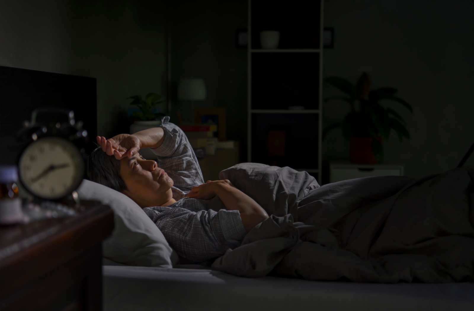 A senior lying awake in bed in the middle of the night stares at the ceiling, holding a hand to their head, waiting for sleep