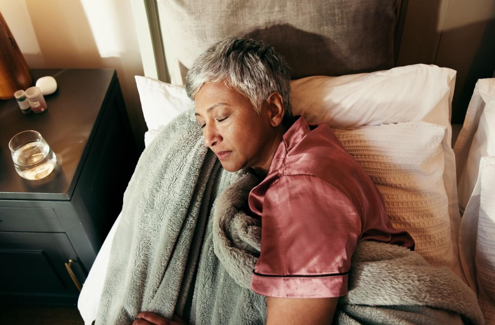 A senior in silk pyjamas sleeps peacefully in a bed full of soft, comforting blankets and extra pillows, with water nearby