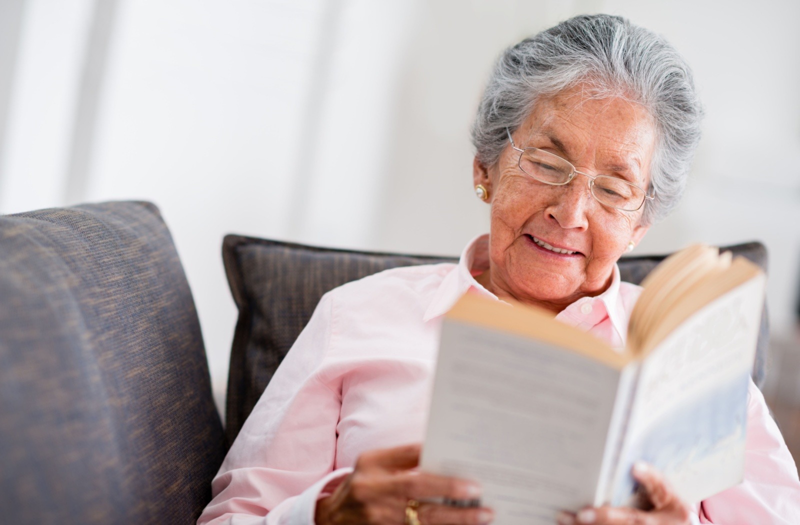 A senior woman reads a paperback novel while sitting on a gray couch.
