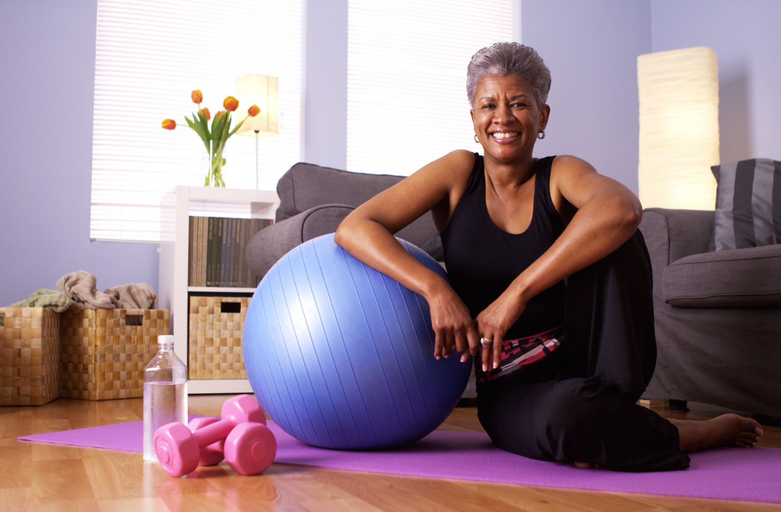 A senior woman poses on a yoga mat while leaning on an inflatable exercise ball with pink dumbbells in the foreground.